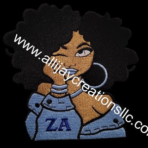 May include: Embroidered patch featuring a stylized Black woman with large black afro, wearing a blue denim top with the letters "ZA". The woman has large silver hoop earrings and blue lipstick. The website address "www.allijaycreationsllc.com" is printed on the patch.