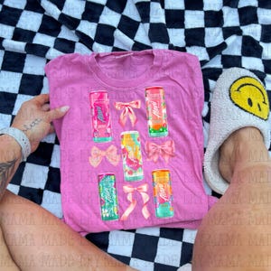 May include: Pink t-shirt featuring colorful Alani Energy drink cans and pink bows. The cans have various fruit designs. The shirt is displayed on a black and white checkered surface.