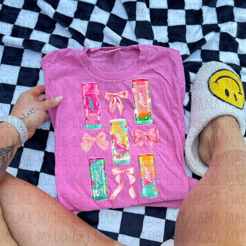May include: Pink t-shirt featuring colorful Alani Energy drink cans and pink bows. The cans have various fruit designs. The shirt is displayed on a black and white checkered surface.
