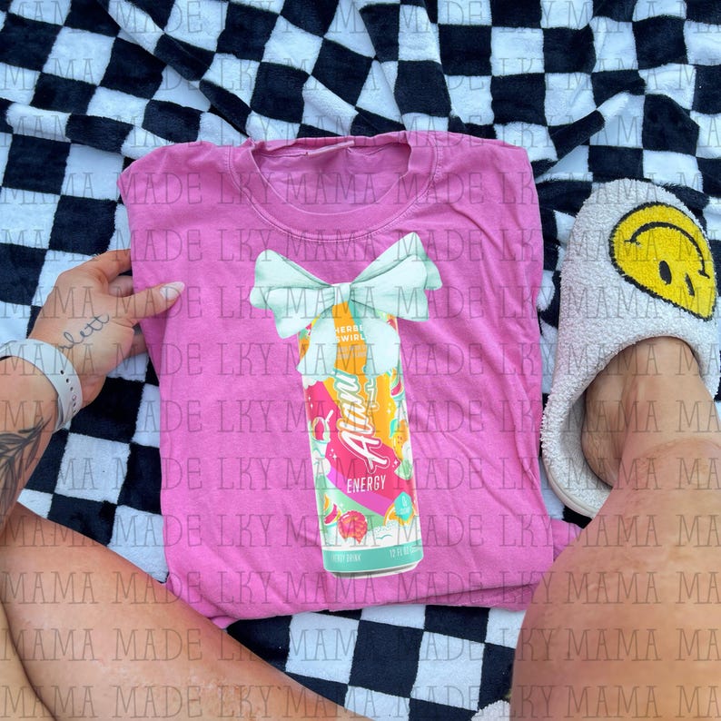May include: A pink t-shirt featuring a graphic of an "Alani Energy" drink can with a bow. The can design includes the words "Cherry Swirl" and "Energy". The shirt is on a black and white checkered blanket.