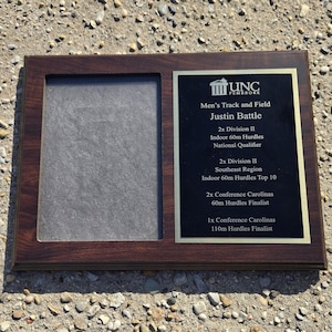 May include: A wooden plaque with a dark brown frame and a black plate. The plaque features text that reads "UNC Pembroke Men's Track and Field Justin Battle" and lists athletic achievements. There is a space for a photo.