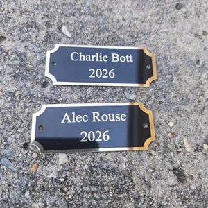 May include: Two black nameplates with gold trim and the names "Charlie Bott 2026" and "Alec Rouse 2026" in white lettering. The nameplates are rectangular with decorative edges and two small holes for mounting.