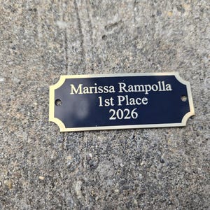 May include: A rectangular, navy blue plaque with gold trim and the text "Marissa Rampolla 1st Place 2026". The plaque has two small holes for mounting and is set against a textured gray surface.