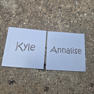 May include: Two light blue square cards with the names "Kyle" and "Annalise" printed in a handwritten style. The cards are placed on a rough, gray concrete surface.
