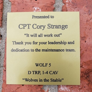 May include: A gold-colored plaque with black text, mounted on a red brick wall. The plaque reads "Presented to CPT Cory Strange" and includes a quote, a thank you message, and unit information. The plaque is rectangular.