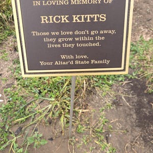 May include: A rectangular bronze memorial plaque with a raised gold border and gold lettering. The text reads: IN LOVING MEMORY OF RICK KITTS. Those we love don't go away, they grow within the lives they touched. With love, Your Altar'd State Family.