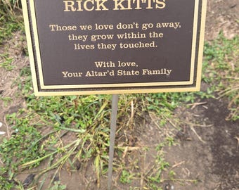 May include: A rectangular bronze memorial plaque with a raised gold border and gold lettering. The text reads: IN LOVING MEMORY OF RICK KITTS. Those we love don't go away, they grow within the lives they touched. With love, Your Altar'd State Family.