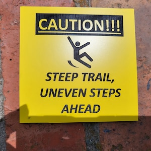 May include: Yellow caution sign with black text and a falling person graphic. The sign reads "CAUTION!!! STEEP TRAIL, UNEVEN STEPS AHEAD" and is mounted on a red brick wall. The sign warns of potential hazards.