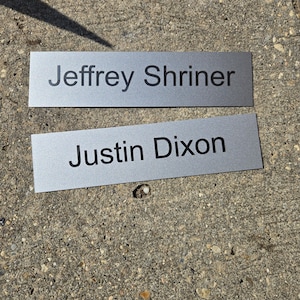 May include: Two rectangular silver nameplates with a brushed metallic finish. The top plate features the black printed text Jeffrey Shriner, and the bottom plate features the black printed text Justin Dixon in a clean, sans-serif font style.