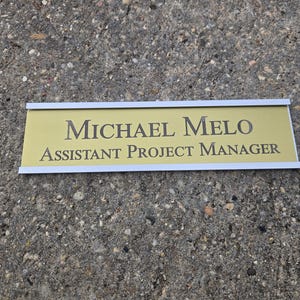 May include: A gold-colored nameplate with the name "MICHAEL MELO" and the title "ASSISTANT PROJECT MANAGER" in black lettering. The nameplate has a white border and is set against a textured gray surface.