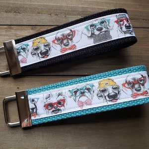 May include: Two keychains with black and turquoise fabric straps. Each strap features a white background with a repeating pattern of dogs wearing glasses. The dogs are wearing hats and bowties.
