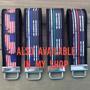 Op de afbeelding: Vier sleutelhangers met zwarte banden en zilveren sluitingen. Elke sleutelhanger heeft een ander Amerikaans vlagontwerp met een dunne rode, blauwe of groene streep. De tekst "ALSO AVAILABLE IN MY SHOP" is zichtbaar in de afbeelding.