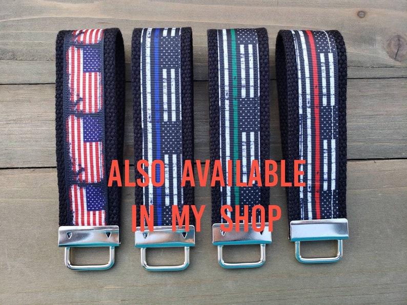 Op de afbeelding: Vier sleutelhangers met zwarte banden en zilveren sluitingen. Elke sleutelhanger heeft een ander Amerikaans vlagontwerp met een dunne rode, blauwe of groene streep. De tekst "ALSO AVAILABLE IN MY SHOP" is zichtbaar in de afbeelding.