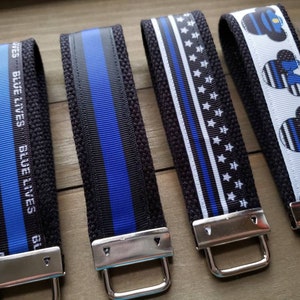 Op de afbeelding: Vier zwarte en blauwe sleutelhangers met zilveren metalen ringen. De sleutelhangers hebben een dunne blauwe lijn, een blauwe lijn met sterren, een blauwe lijn met de tekst "Blue Lives Matter" en een Mickey Mouse-ontwerp met een blauwe lijn.