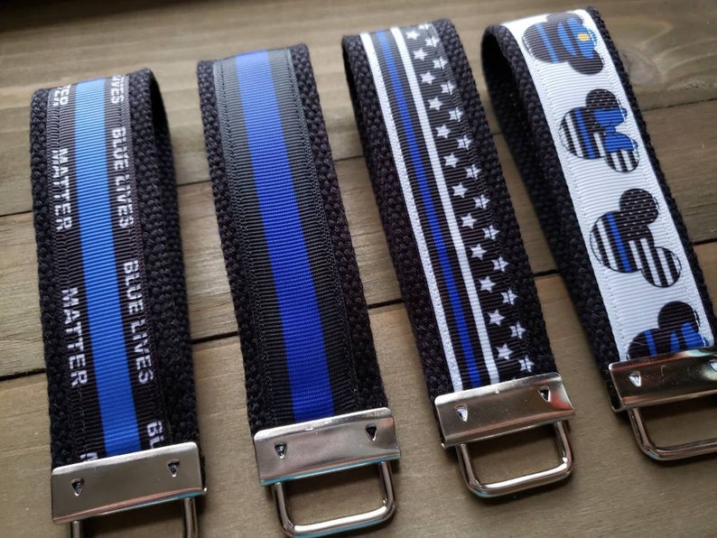 Op de afbeelding: Vier zwarte en blauwe sleutelhangers met zilveren metalen ringen. De sleutelhangers hebben een dunne blauwe lijn, een blauwe lijn met sterren, een blauwe lijn met de tekst "Blue Lives Matter" en een Mickey Mouse-ontwerp met een blauwe lijn.