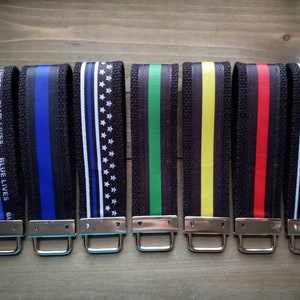 Op de afbeelding: Zeven zwarte sleutelhangers met verschillende gekleurde lintontwerpen. De linten omvatten blauw en zwart, witte en zwarte sterren, groen en zwart, geel en zwart, rood en zwart en witte en zwarte strepen. De sleutelhangers hebben zilveren metalen ringen.