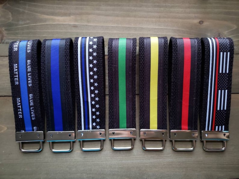 Op de afbeelding: Zeven zwarte sleutelhangers met verschillende gekleurde lintontwerpen. De linten omvatten blauw en zwart, witte en zwarte sterren, groen en zwart, geel en zwart, rood en zwart en witte en zwarte strepen. De sleutelhangers hebben zilveren metalen ringen.