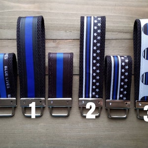 Op de afbeelding: Vijf verschillende sleutelhangers met blauwe en zwarte lintontwerpen. De eerste sleutelhanger heeft een blauw en zwart lint met de tekst "Blue Lives Matter". De tweede sleutelhanger heeft een blauw en zwart lint. De derde sleutelhanger heeft een wit en zwart lint met blauwe sterren. De vierde sleutelhanger heeft een wit lint met blauwe sterren en een zwart en blauw Mickey Mouse-ontwerp. De vijfde sleutelhanger heeft een wit lint met blauwe sterren en een zwart en blauw Mickey Mouse-ontwerp.