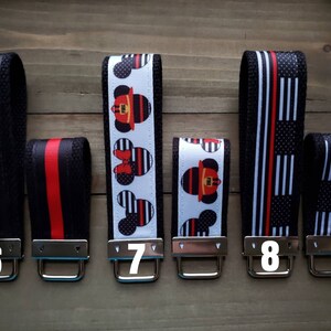 Op de afbeelding: Vijf sleutelhangers met verschillende ontwerpen. De sleutelhangers zijn zwart met rode en witte strepen. De ontwerpen omvatten een brandweerman Mickey Mouse, een rood-wit gestreept ontwerp en een zwart-wit gestreept ontwerp. De sleutelhangers zijn genummerd 6, 7, 8.