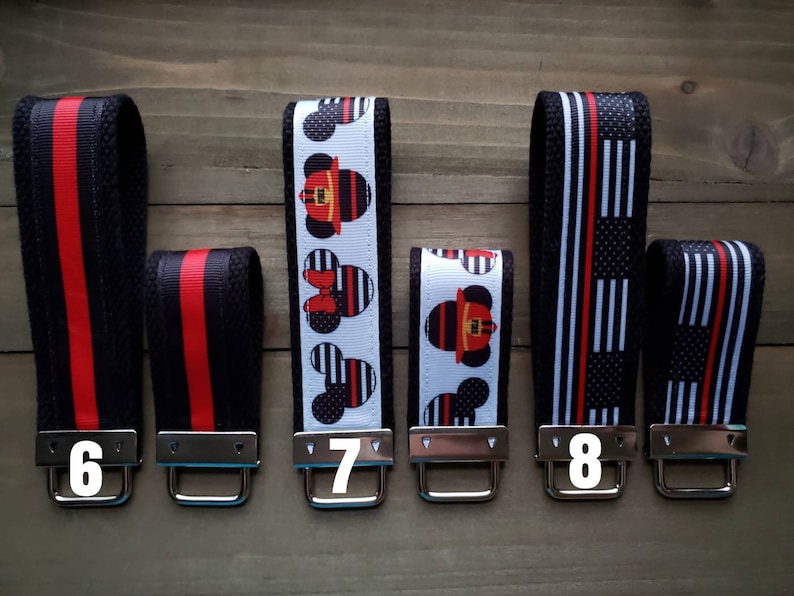 Op de afbeelding: Vijf sleutelhangers met verschillende ontwerpen. De sleutelhangers zijn zwart met rode en witte strepen. De ontwerpen omvatten een brandweerman Mickey Mouse, een rood-wit gestreept ontwerp en een zwart-wit gestreept ontwerp. De sleutelhangers zijn genummerd 6, 7, 8.