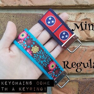 Op de afbeelding: Twee sleutelhangers, een met een bloemenpatroon op een turquoise achtergrond en de andere met de vlag van de staat Tennessee op een blauwe achtergrond. Beide sleutelhangers hebben zilveren sleutelringen. De tekst "Mini" en "Regular" staat op de bakstenen muur achter de sleutelhangers. De tekst "ALL KEYCHAINS COME WITH A KEYRING!" staat onder de sleutelhangers.