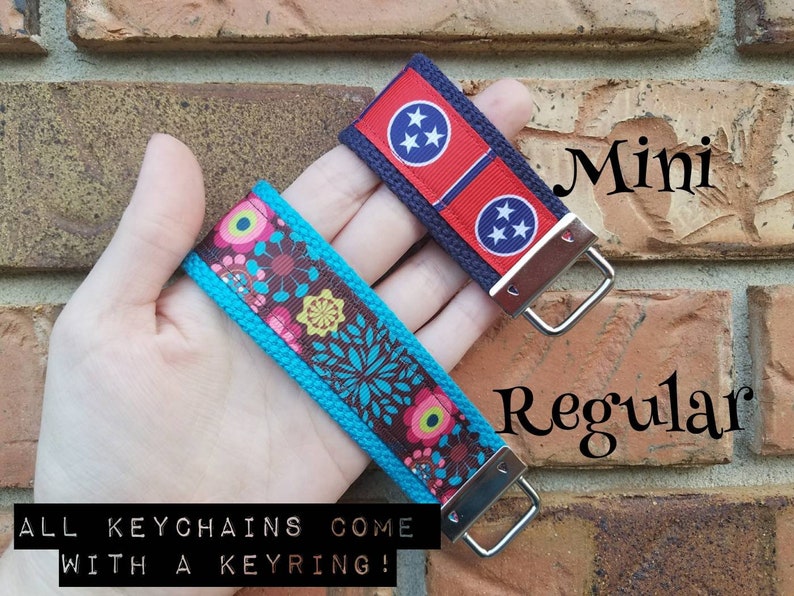 Op de afbeelding: Twee sleutelhangers, een met een bloemenpatroon op een turquoise achtergrond en de andere met de vlag van de staat Tennessee op een blauwe achtergrond. Beide sleutelhangers hebben zilveren sleutelringen. De tekst "Mini" en "Regular" staat op de bakstenen muur achter de sleutelhangers. De tekst "ALL KEYCHAINS COME WITH A KEYRING!" staat onder de sleutelhangers.