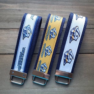 May include: Three keychains with a blue, white, and yellow design featuring the Nashville Predators logo. The keychains are made of fabric and have a silver metal ring.