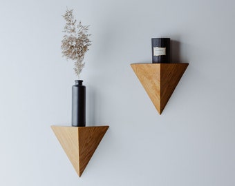 May include: Two triangular wooden shelves mounted on a white wall. The top shelf has a black candle on it. The bottom shelf has a black vase with dried flowers.