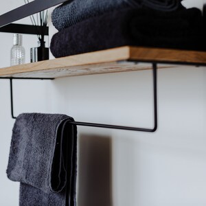 May include: A black metal shelf with a wooden top and a towel bar underneath. The shelf is mounted to a white wall and has a few towels and bottles on it.