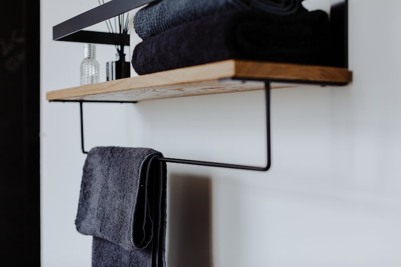 May include: A black metal shelf with a wooden top and a towel bar underneath. The shelf is mounted to a white wall and has a few towels and bottles on it.