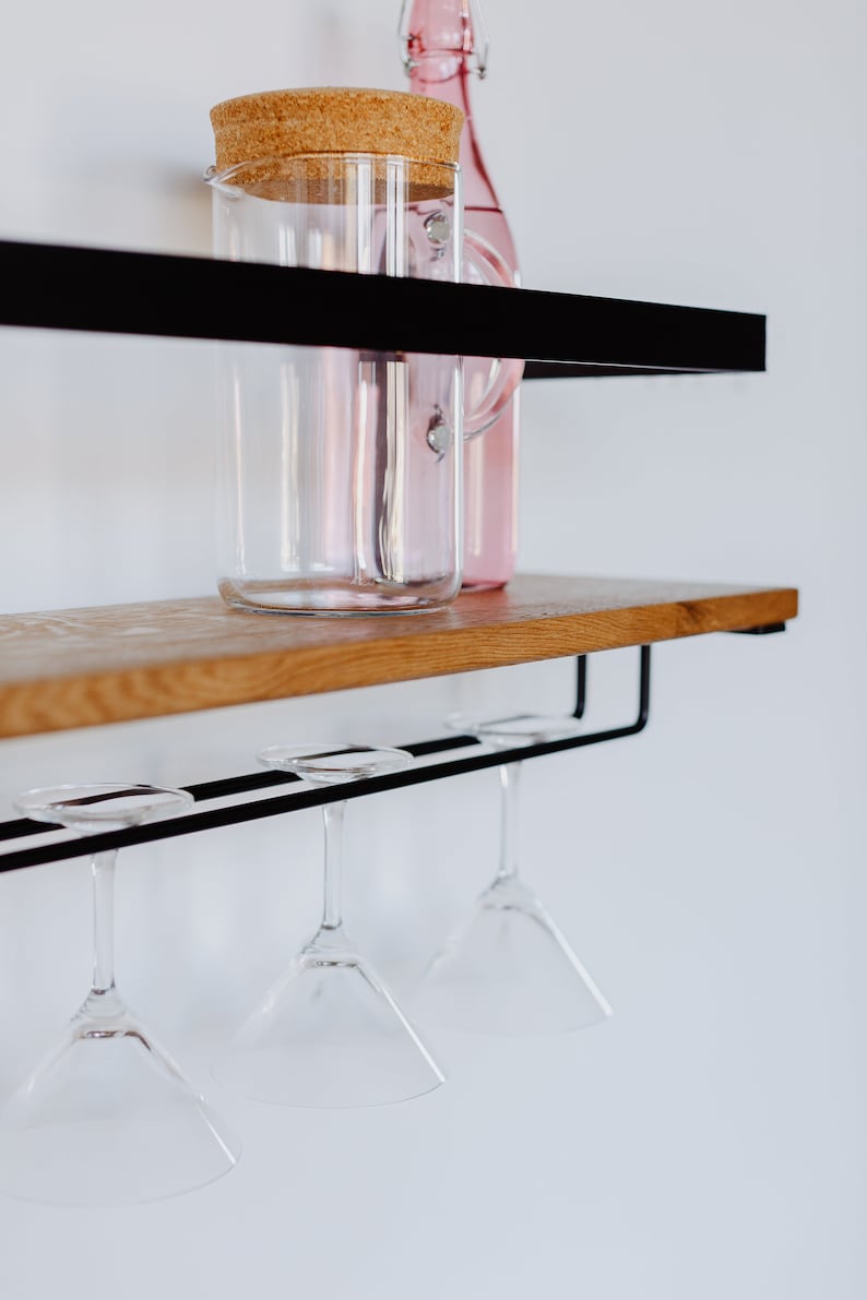 May include: A black metal shelf with a wooden top, holding a glass pitcher with a cork lid and a pink glass bottle.  Four martini glasses hang from the bottom shelf.