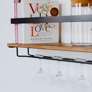 May include: A wooden shelf with a black metal frame and a glass jar with a cork lid. There are four wine glasses hanging from the shelf and a stack of cookbooks with the title "Vege Love" by Barbara Kafka.