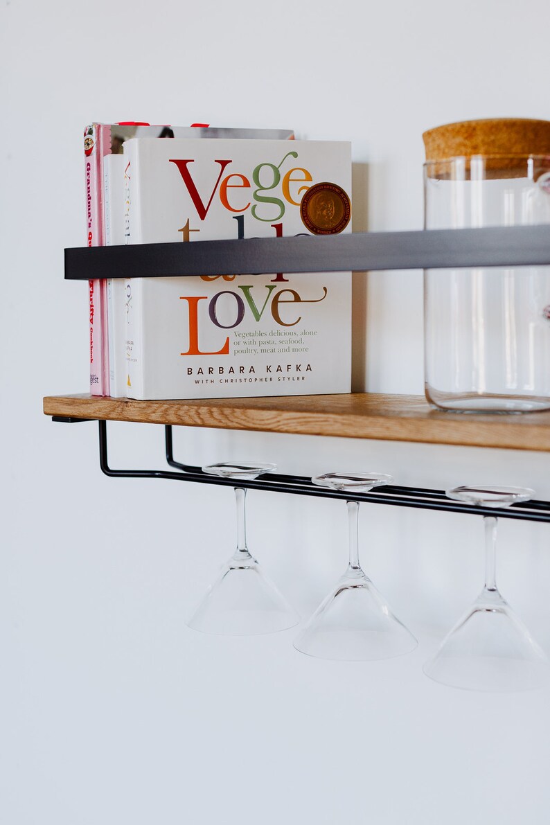 May include: A wooden shelf with a black metal frame and a glass jar with a cork lid. There are four wine glasses hanging from the shelf and a stack of cookbooks with the title "Vege Love" by Barbara Kafka.