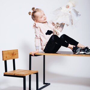 May include: A young girl sits on a wooden table with black metal legs, holding a white doll with wings. A small wooden chair with black metal legs sits in the foreground.