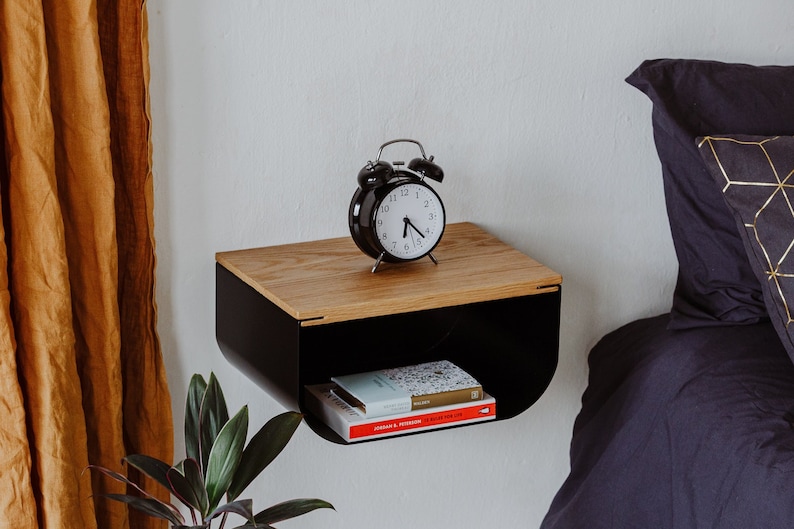 May include: A black metal wall shelf with a wood top. The shelf has a curved front and is mounted on a white wall. A black alarm clock and a stack of books are on the shelf.