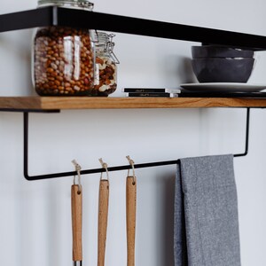 May include: A black metal shelf with a wooden top, featuring two glass jars filled with nuts and beans. The shelf has a black metal bar with hooks underneath, holding a gray kitchen towel and three wooden-handled kitchen utensils.