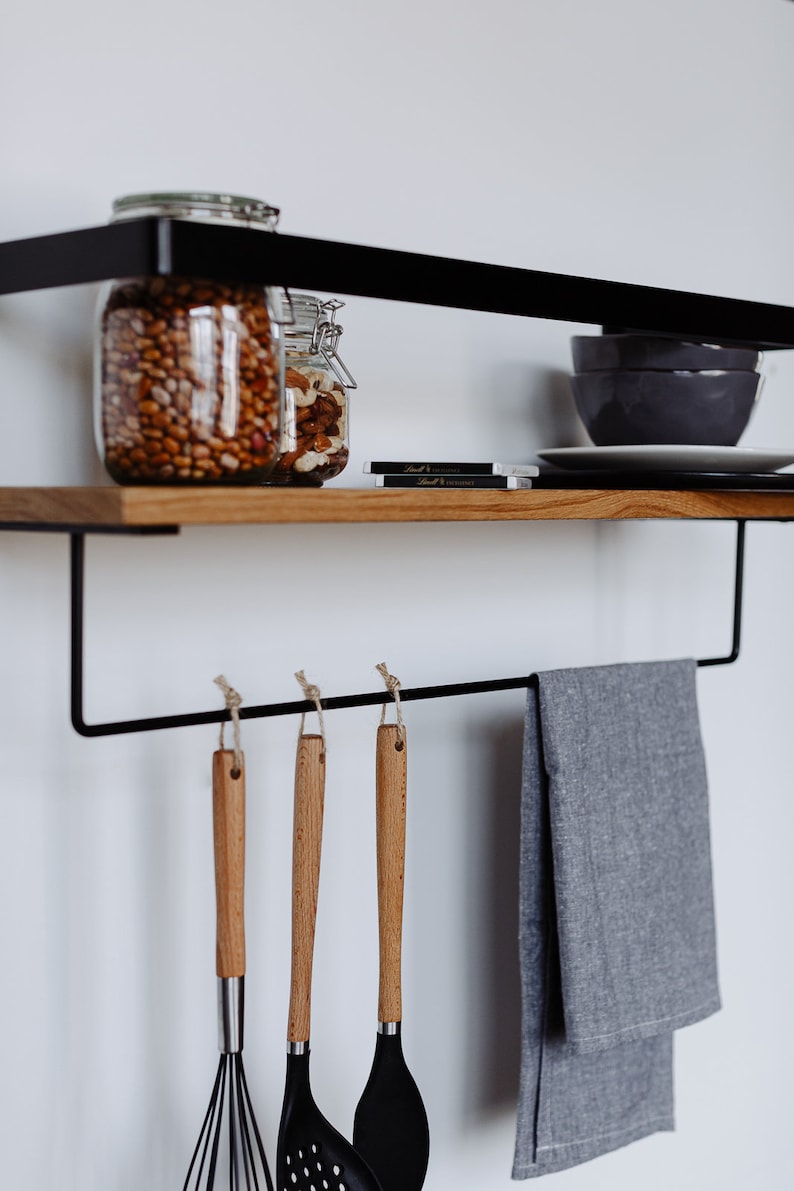 May include: A black metal shelf with a wooden top, featuring two glass jars filled with nuts and beans. The shelf has a black metal bar with hooks underneath, holding a gray kitchen towel and three wooden-handled kitchen utensils.