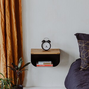 May include: A black floating shelf with a wooden top, a black alarm clock, and a stack of books. The shelf is mounted on a white wall next to a bed with a black duvet and a gold patterned pillow.
