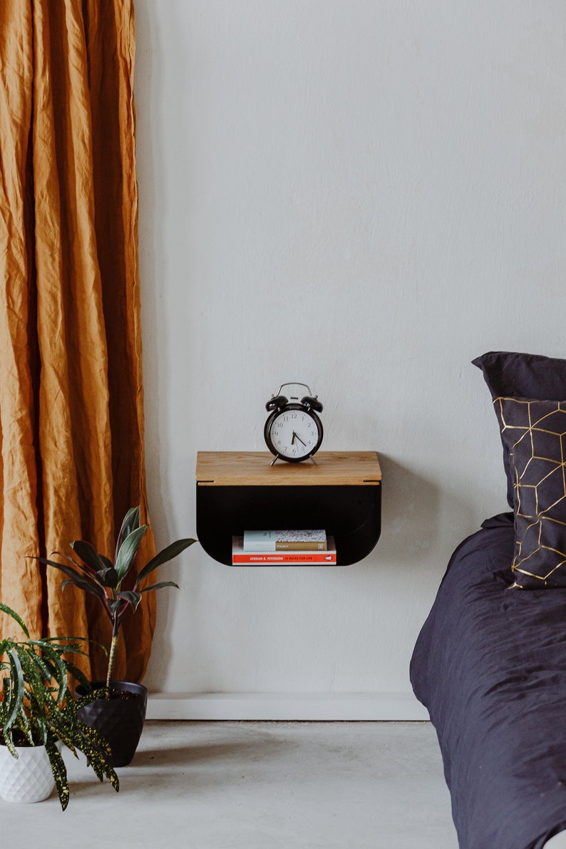 May include: A black floating shelf with a wooden top, a black alarm clock, and a stack of books. The shelf is mounted on a white wall next to a bed with a black duvet and a gold patterned pillow.