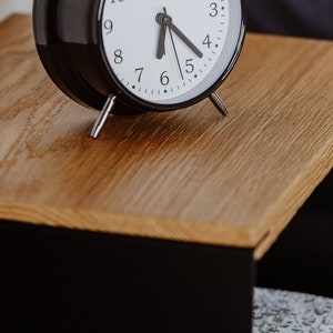 May include: A black alarm clock with white face and black hands set to 5:05 on a wooden surface.