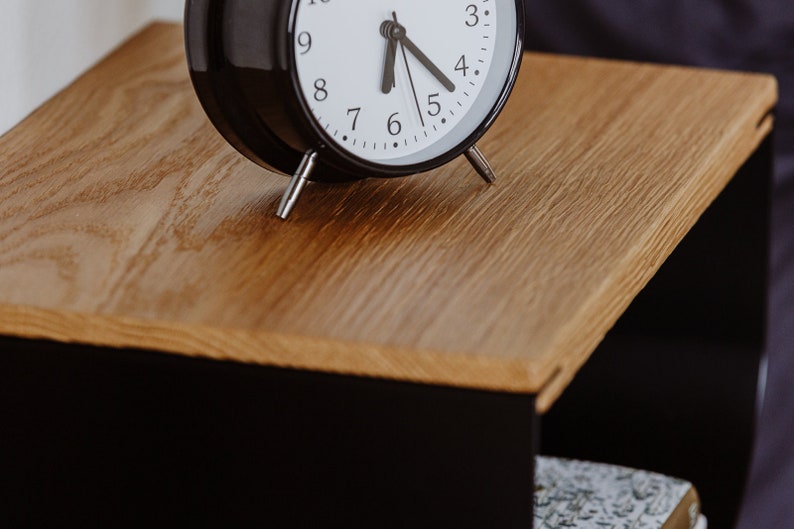 May include: A black alarm clock with white face and black hands set to 5:05 on a wooden surface.