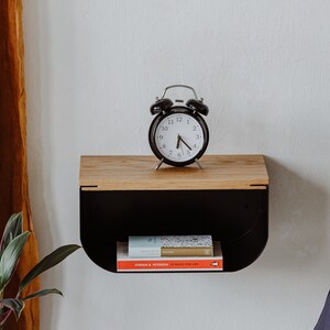 May include: A black alarm clock with white face and black hands sits on a wooden shelf with a black metal frame. Two books are on the shelf below the clock.