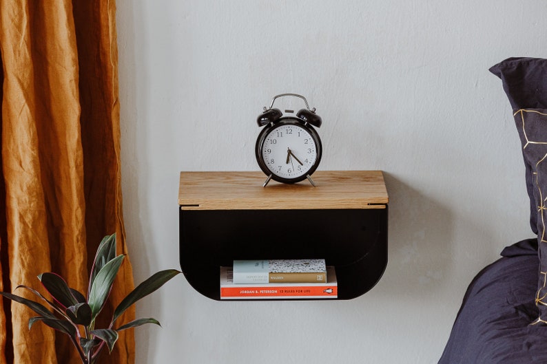 May include: A black alarm clock with white face and black hands sits on a wooden shelf with a black metal frame. Two books are on the shelf below the clock.