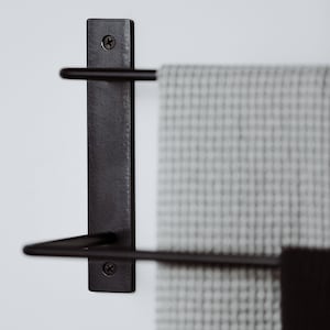 Pode incluir: Toalheiro de metal preto com duas barras montadas em uma parede branca. Uma toalha cinza est&aacute; pendurada no toalheiro.