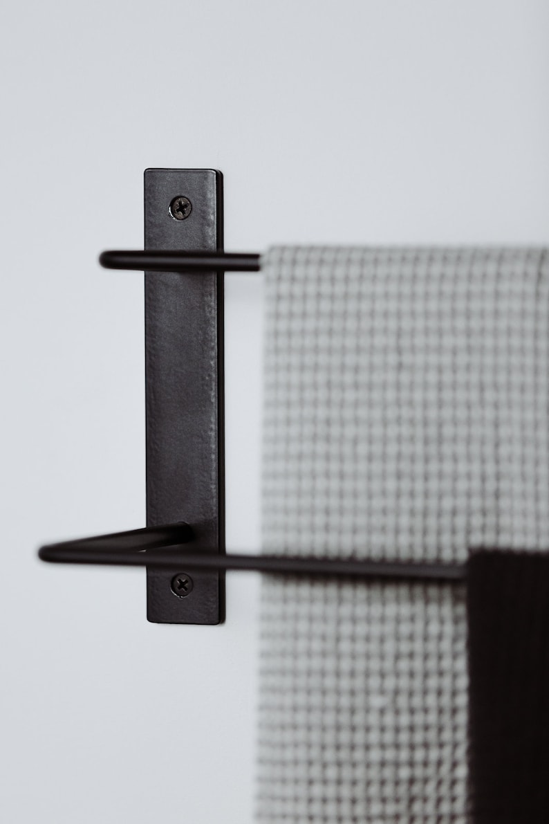 Pode incluir: Toalheiro de metal preto com duas barras montadas em uma parede branca. Uma toalha cinza est&aacute; pendurada no toalheiro.