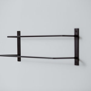 Pode incluir: Um suporte de toalha de parede de metal preto com duas barras. O suporte &eacute; montado em uma parede branca.