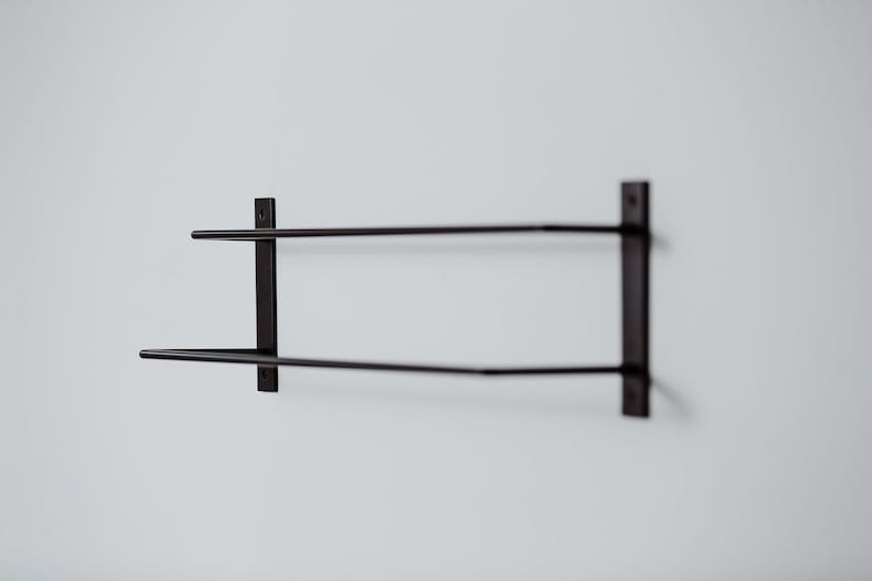Pode incluir: Um suporte de toalha de parede de metal preto com duas barras. O suporte &eacute; montado em uma parede branca.
