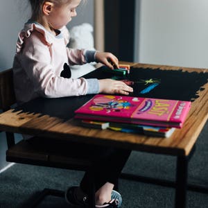 May include: A young girl sits at a wooden table with a black chalkboard top. She is drawing with colorful chalk and has a pink book in front of her.