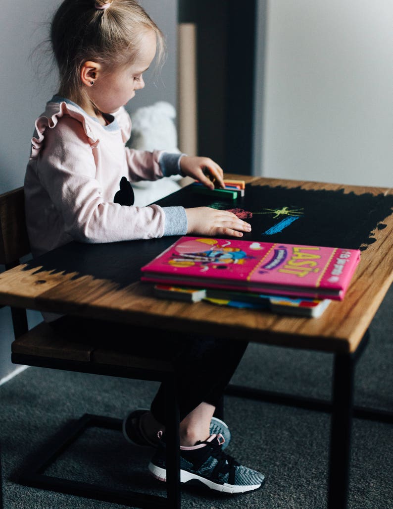May include: A young girl sits at a wooden table with a black chalkboard top. She is drawing with colorful chalk and has a pink book in front of her.