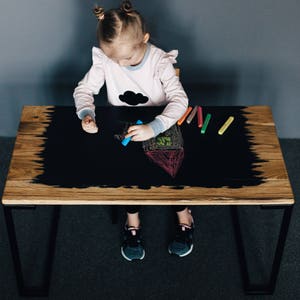 May include: A young child sits at a black chalkboard table drawing with colorful chalk. The table has a wooden top and black metal legs.